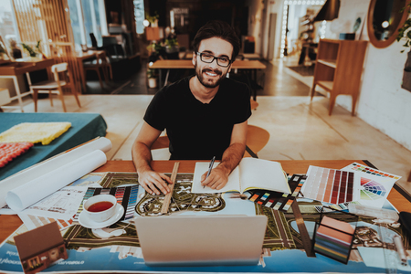 Portrait of Smiling Graphic Designer Enjoys Work. Handsome Caucasian Bearded Illustrator Looking at Camera Sits at Table Doing Creative Project at Workplace. Dream Work Conceptの写真素材