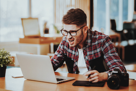 Nervous Photographer Works on Laptop Remotely. Portrait of Serious Stylish Freelance Graphic Designer Sits at Table Retouch Photo Frowns While Looking at Screen Indoors. Distance Job Conceptの写真素材