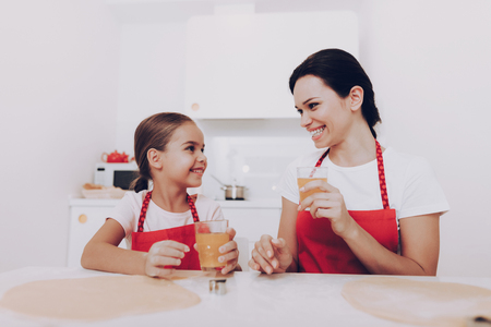 Mom and Young Daughter Drinking Sweet Juice. Apron on Mom and Daughter when Cook. Smile Mom and Daughter. Happy Family Together. Beautiful Girl with Mom Preparation for Cook. Happy Girl with Mom.の写真素材