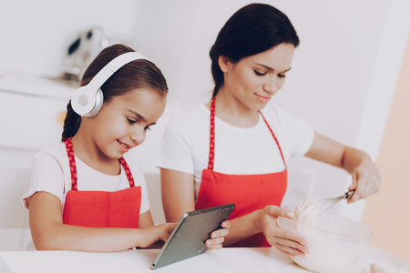 Girl with Headphones Looks How Bake a Cake. Love Mother Help Daughter with Bake. Dother Looks How do it. Happy Girl with Young Mother Bace Good Breakfast. Little Girl Cook Food with Mother in Kitchen.の写真素材