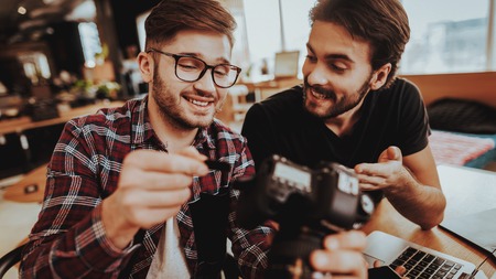 Closeup of Smiling Guy Insert SD Card in Camera. Portrait of Two Smiling Handsome Bearded Photographers Plug SD Card In Camera Slot. Media Data Transferring and Storage Device Conceptの写真素材