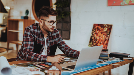 Portrait of Young Graphic Designer Work on Laptop Indoors. Bearded Illustrator Wearing Glasses Sitting at Workplace Doing Design Project Looking at PC Screen. Creative Job Conceptの写真素材