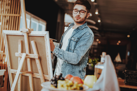 Portrait of Guy Painting on Easel Still Life. Handsome Professional Caucasian Talanted Painter Artist in Casual Clothes Looking at Fuits Standing in Bright Room. Art Educationの写真素材