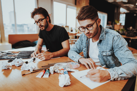Two Talanted Freelance Artists Drawing Indoors. Portrait of Painters Sitting at Table Focused on Art Project Using Paintbrush and Pencils Coworking in Bright Room. Cooperation Conceptの写真素材