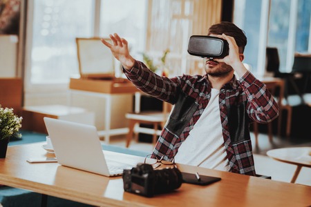 Happy Freelancer Working Remotely and Testing VR. Portrait of Cheerful Caucasian Guy Wears Virtual Reality Goggles Sitting at Desk Using Laptop ang Gadgets Holds Hand Up Indoors.の写真素材