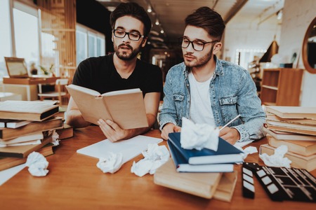 Two Concentrated Text Writers Working Together Indoors. Handsome Screenwriters Scenarist Sitting at Table Near Stacks of Books Reading Textbook, Novel, Play . Work Collaboration Conceptの写真素材