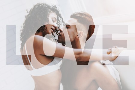 Young Beautiful Couple. Couple is Caucasian Boy and Black Curly Girl. Couple is Sitting on Bed. People is Kissing and Feeling a Good. Sexual People is Naked. Sexual Prelude. People Located in Bedroom.の写真素材