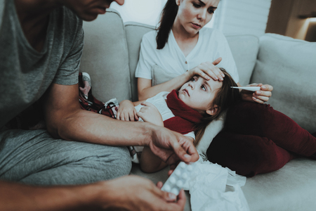 Little Girl in Red Scarf with Cold with Family. Sick Young Girl. White Sofa in Room. Father and Mother. Disease Concept. Healthcare and Healthy Lifestyle Concept. Take Care of Daughter.の写真素材