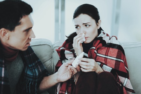Sick Couple Sitting on Sofa in Checkered Blankets. Man at Home. Sick Young Woman. White Sofa in Room. Unhappy Guy. Disease Concept. Healthcare and Healthy Lifestyle Concept. Using Nasal Spray.の写真素材