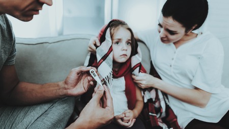 Little Girl in Red Scarf with Cold with Family. Sick Young Girl. White Sofa in Room. Father and Mother. Disease Concept. Healthcare and Healthy Lifestyle Concept. Take Care of Daughter.の写真素材