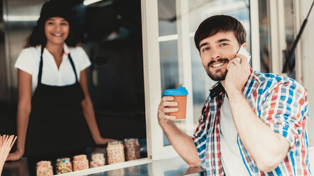 Young Man in Shirt Buying Coffee in Food Truck. Promenade in Town. Summer Day. Guy in Checkered Shirt. Street Food Concept. Cup of Coffee. Buying Food Outdoor. Food in Town. Talking on Smartphone.の写真素材
