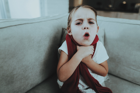 Little Girl with Cold Sitting on Sofa in Red Scarf. Sick Young Girl. White Sofa in Room. Unhappy Child. Disease Concept. Healthcare and Healthy Lifestyle Concept. Little Girl at Home.の写真素材