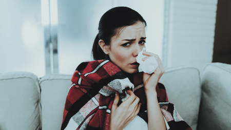 Woman with Cold on Sofa in Checkered Blanket. Girl at Home. Sick Young Woman. White Sofa in Room. Unhappy Woman. Disease Concept. Healthcare and Healthy Lifestyle Concept. Using Napkins.の写真素材