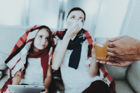Sick Family Sitting on Sofa in Checkered Blankets. Mother and Daughter. White Sofa in Room. Unhappy Girl. Disease Concept. Healthcare and Healthy Lifestyle Concept. Using Serviettes. Girl with Laptop.の写真素材
