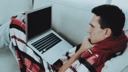 Man with Cold Sitting on Sofa in Checkered Blanket. Man at Home. Sick Young Man. White Sofa in Room. Unhappy Guy. Disease Concept. Healthcare and Healthy Lifestyle Concept. Using Laptop.の写真素材