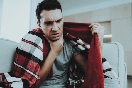Man with Cold Sitting on Sofa in Checkered Blanket. Man at Home. Sick Young Man. White Sofa in Room. Unhappy Guy. Disease Concept. Healthcare and Healthy Lifestyle Concept. Man in Red Scarf.の写真素材