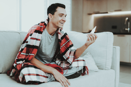 Man with Cold Sitting on Sofa in Checkered Blanket. Man at Home. Sick Young Man. White Sofa in Room. Disease Concept. Healthcare and Healthy Lifestyle Concept. Using Spray. Flu Treatment.の写真素材