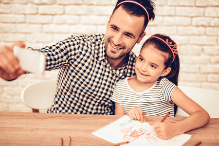 Father and Daughter on Sofa. Spend Time Together. Father's day. White Interior. Headband Hair on Head. Cakes on Plates. Bottle with Topping in Hand. Draw Picture on Table. Smiling Person Take Selfie.の写真素材