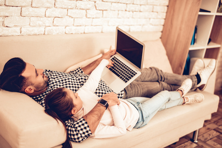 Sit on Sofa. Laptop in Hand. Father and Daughter in Apartment. Spend Time Together. Father's day. Man Playing with Girl. White Interior. Lying on Couch with Laptop. Watch Movie in Laptop.の写真素材