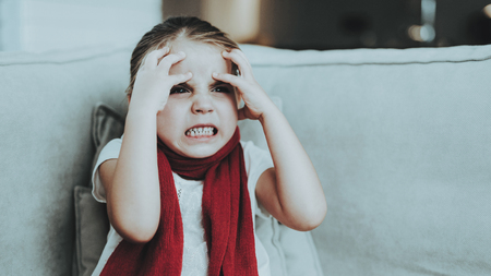 Little Girl with Cold Sitting on Sofa in Red Scarf. Sick Young Girl. White Sofa in Room. Unhappy Child. Disease Concept. Healthcare and Healthy Lifestyle Concept. Little Girl at Home.の写真素材