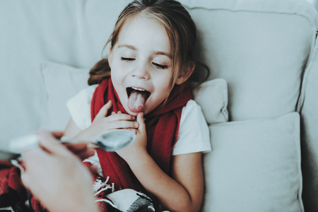 Little Girl with Cold Sitting on Sofa in Red Scarf. Medicinal Syrup. Sick Young Girl. White Sofa in Room. Unhappy Child. Disease Concept. Healthcare and Healthy Lifestyle Concept. Little Girl at Home.の写真素材