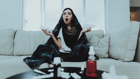 Upset Woman with Cold on Sofa in Scarf at Home. Sick Young Woman. White Sofa in Room. Unhappy Woman. Disease Concept. Healthcare and Healthy Lifestyle Concept. Unhappy Girl. Modern Medicine.の写真素材
