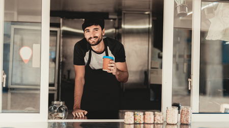 Young Bearded Man in Apron Standing in Food Truck. Cup of Coffee. Street Food Concept. Food in Town. Selling Snacks. Guy in Cap. Black Arpon. Working Outdoor. Man in T-shirt. Standing Guy.の写真素材