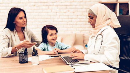 Mother with Son at Arabic Doctor's Appointment. Confident Muslim Female Doctor. Child at the Pediatrician. Hospital Concept. Healthy Concept. Pediatrician Writes a Prescription. Child Patient Visiting Doctor.の写真素材
