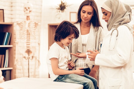 Arabic Doctor Give Medicine to Sick Boy Lying Bed. Confident Muslim Female Doctor Giving Glass of Medicine. Child at the Pediatrician. Hospital Concept. Healthy Concept. Child Patient Visiting Doctor.の写真素材