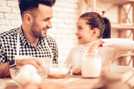 Cook Food at Home. Happy Family. Father's Day. Girl and Man Cook Food. Smiling Man and Child at Table. Spend Time Together. Food on Table. Break Egg in Bowl. Pour Flour. Cook Dough. Pour Flour in Bowlの写真素材