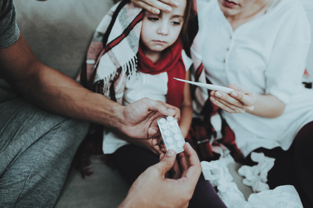 Little Girl in Red Scarf with Cold with Family. Sick Young Girl. White Sofa in Room. Father and Mother. Disease Concept. Healthcare and Healthy Lifestyle Concept. Take Care of Daughter.の写真素材