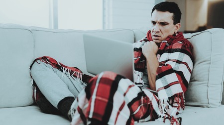 Man with Cold Sitting on Sofa in Checkered Blanket. Man at Home. Sick Young Man. White Sofa in Room. Unhappy Guy. Disease Concept. Healthcare and Healthy Lifestyle Concept. Using Laptop.の写真素材