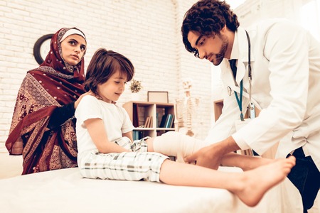 Arabic Male Doctor Bandaging Limb of Child Patient. Hospital Concept. Healthy Concept. Child Patient Visiting Doctor. Doctor Bandaging Leg Little Patien. Child Patient Visiting Doctor. Leg Injuryの写真素材