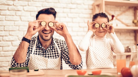 Cook Food at Home. Father Feeds Daughter. Happy Family. Father's Day. Girl and Man Cook Food. Spend Time Together. Sliced Tomatoes on Board. Man with Vegetables. Sliced Cucumbers near Eyes.の写真素材
