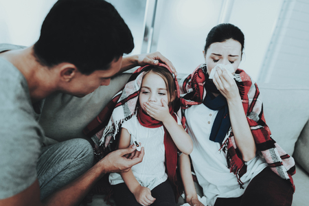 Sick Family Sitting on Sofa in Checkered Blankets. Mother and Daughter. White Sofa in Room. Unhappy Girl. Disease Concept. Healthcare and Healthy Lifestyle Concept. Using Serviettes. Father with Family.の写真素材