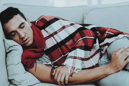 Man with Cold Resting on Sofa in Checkered Blanket. Man at Home. Sick Young Man. White Sofa in Room. Unhappy Guy. Disease Concept. Healthcare and Healthy Lifestyle Concept. Sleeping Guy.の写真素材