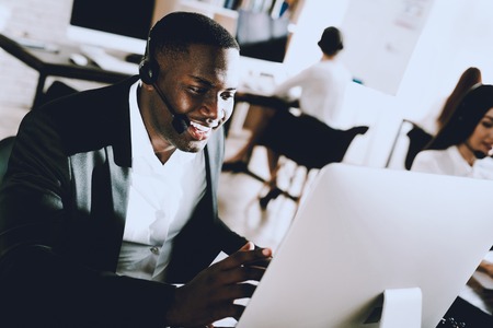 Sitting Afro American Man Working in Call Center. Manager with Computer. Man on Workplace. Consultant with Microphone. Customer Service Concept. Woman in Headphones. Operator in Office.の写真素材