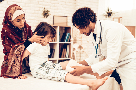 Arabic Male Doctor Bandaging Limb of Child Patient. Hospital Concept. Healthy Concept. Child Patient Visiting Doctor. Doctor Bandaging Leg Little Patien. Child Patient Visiting Doctor. Leg Injuryの写真素材