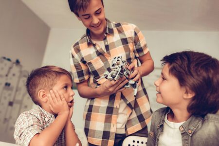 Teenager Demonstrating Robot to Two Sitting Boys. Young Boy in Shirt. Indoor Joy. Modern Hobby Concept. Modern Technology. Robot Engineering Concept. Teenager with Robor. Innovation for Fun.の写真素材