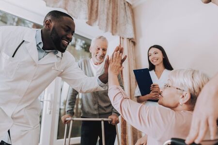 Doctor High Five Patient. Male Rehabilitation. Doctor in Nursing Home. Patient with Walker. Nursing Home. Man with Crutches. Patient on a Wheelchair. Sitting Elderly Woman. Patient Smiling.の写真素材
