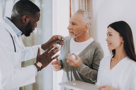 Doctor Tell Nurse about Cure. Elderly Male Listen. Doctor Tell about how Take Pills. Man with Crutches. Nurse Hold Glass of Water. Nursing Home.の写真素材