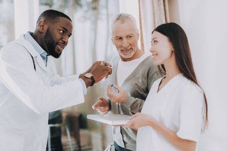 Man with Crutches. Nurse Hold Glass of Water. Doctor Tell Nurse about Cure. Elderly Male Listen. Doctor Tell about how Take Pills. Nursing Home. Patient with Walker. Patient in Rehabilitation.の写真素材