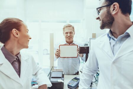 Pharmacists Working at Counter. Customer Holding a Placard. Pharmacists Looking at Customer. Pharmacists Wearing a Special Medical Uniform.の写真素材