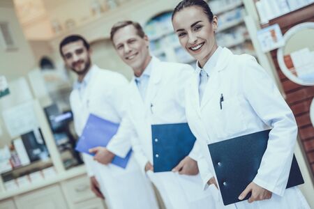 Three Pharmacists Posing. Pharmacists is a Caucasian Men and Young Woman. Persons Holding a Folders. People Wearing a Special Medical Uniform. People is Smiling. People Located in Pharmacy.の写真素材