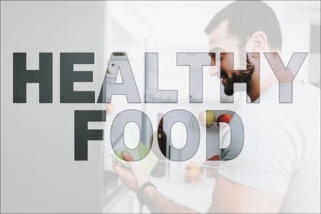 Healthy Food. Athletic Man Posing in the Kitchen. Man Looking in His Refrigerator. Refrigerator with Fresh Vegetables and Fruits. Man Holding a Plastic Container. Man Have a Healthy Lifestyle.の写真素材