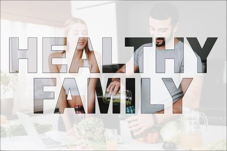 Healthy Family. Athletic Couple in Kitchen. Fresh Vegetables and Fruits on Table. Laptop on Table. Man Pours a Vegetable Cocktail. Girl Looking on a Man. Couple Have a Healthy Lifestyle.の写真素材