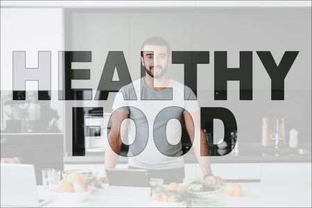 Healthy Food. Man Posing in the Kitchen. Food on Table. Food is Vegetables and Fruits. Laptop Computer on Table. Man Have a Healthy Lifestyle. Man Smiling into Camera.の写真素材