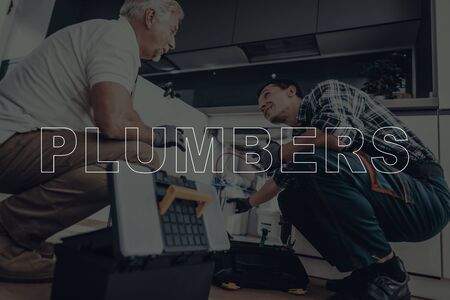 Two Male Plumbers are Working. Plumbers is Adult Guy and Old Man. Workers in a Modern Kitchen. Men Wearing in a Special Uniform. Men Choose the Right Tool for Repairing Equipment. Tool Box on Floor.の写真素材