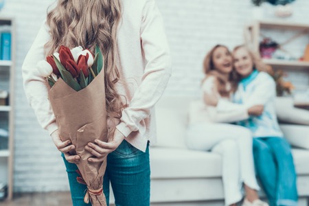 Girl Presenting Bouquet of Tulips to Smiling Mother. Mother with Daughter. Smiling Women. Celebration Concept. Happy Family. Sitting at Home. Red Flower. White Flower. Holiday in March. Mother's Day.の写真素材