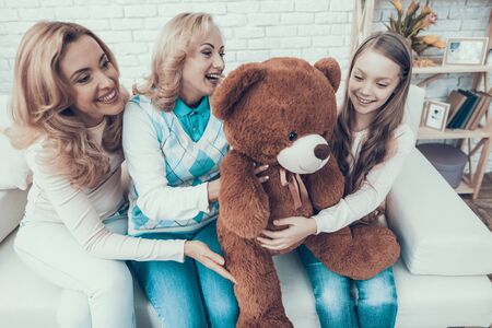 Family Presenting Toy Bear to Smiling Daughter. Gift on Birthday. Mother with Daughter. Smiling Women. Sitting on Sofa. Celebration Concept. Happy Family. Sitting at Home. Holiday in March.の写真素材